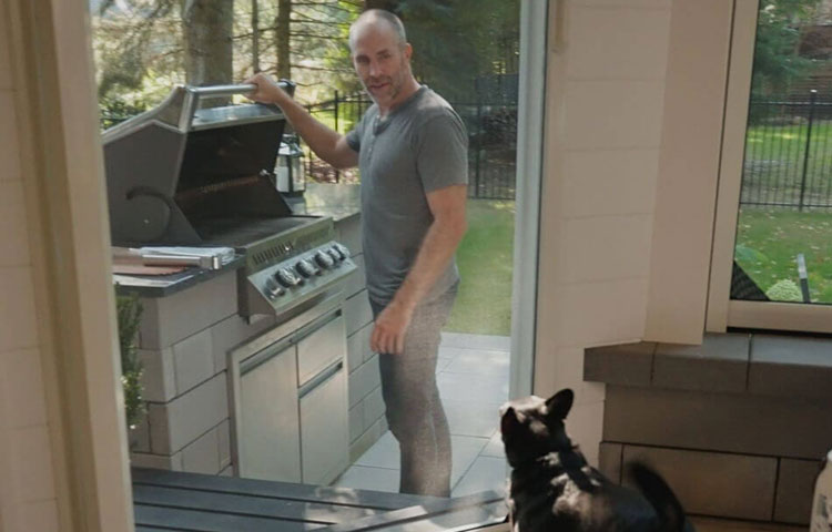 Small dog standing inside looking out through screen door at person grilling