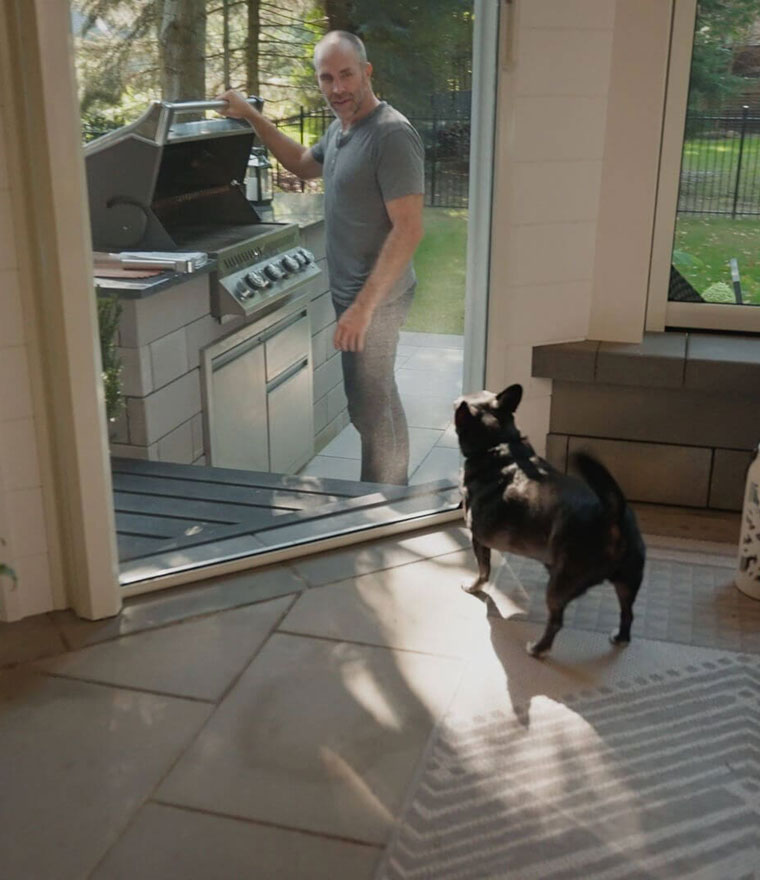 Small dog standing inside looking out through screen door at person grilling